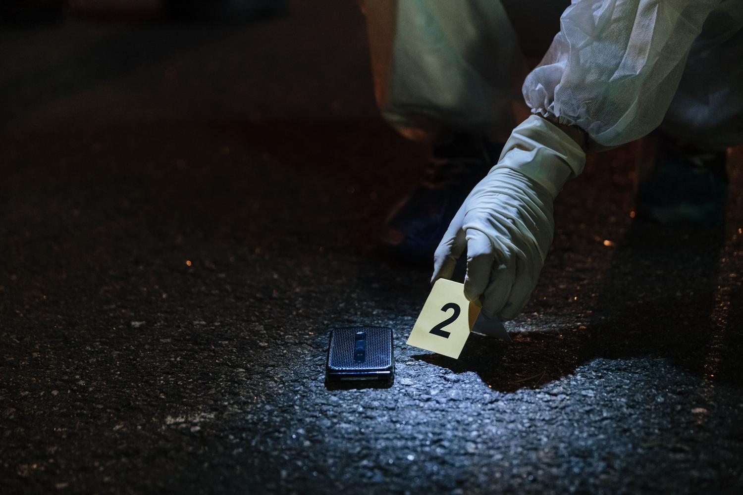 Perito forense recolectando un teléfono móvil como evidencia en una escena del crimen nocturna, utilizando guantes y marcadores.