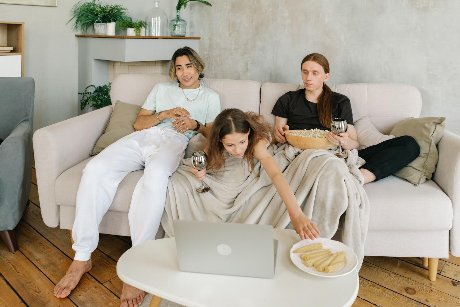 Una familia relajada en el sofá viendo anime juntos en un portátil mientras comparten aperitivos.