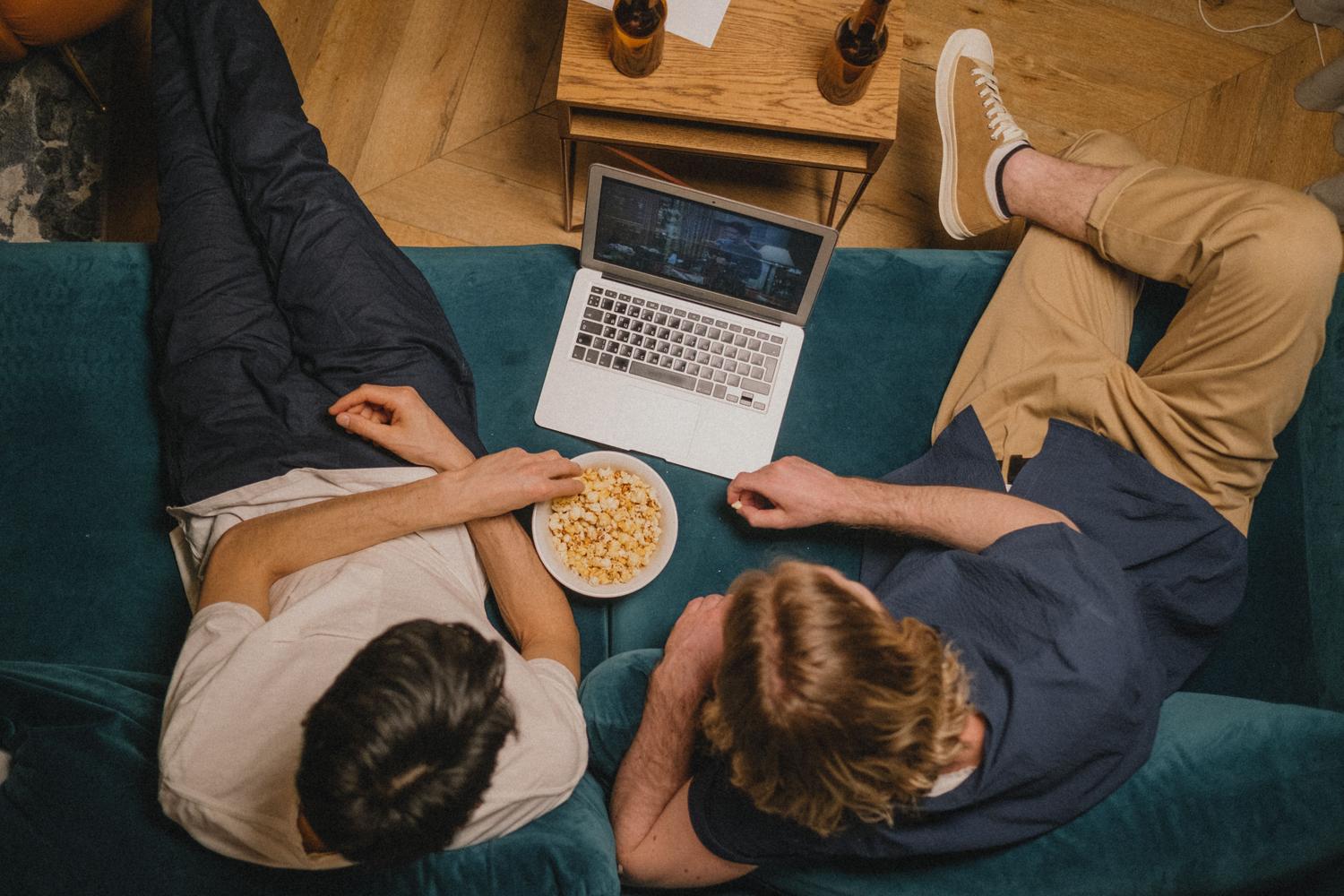 Dos amigos disfrutando de una serie de anime en un ordenador portátil con palomitas en un sofá acogedor.
