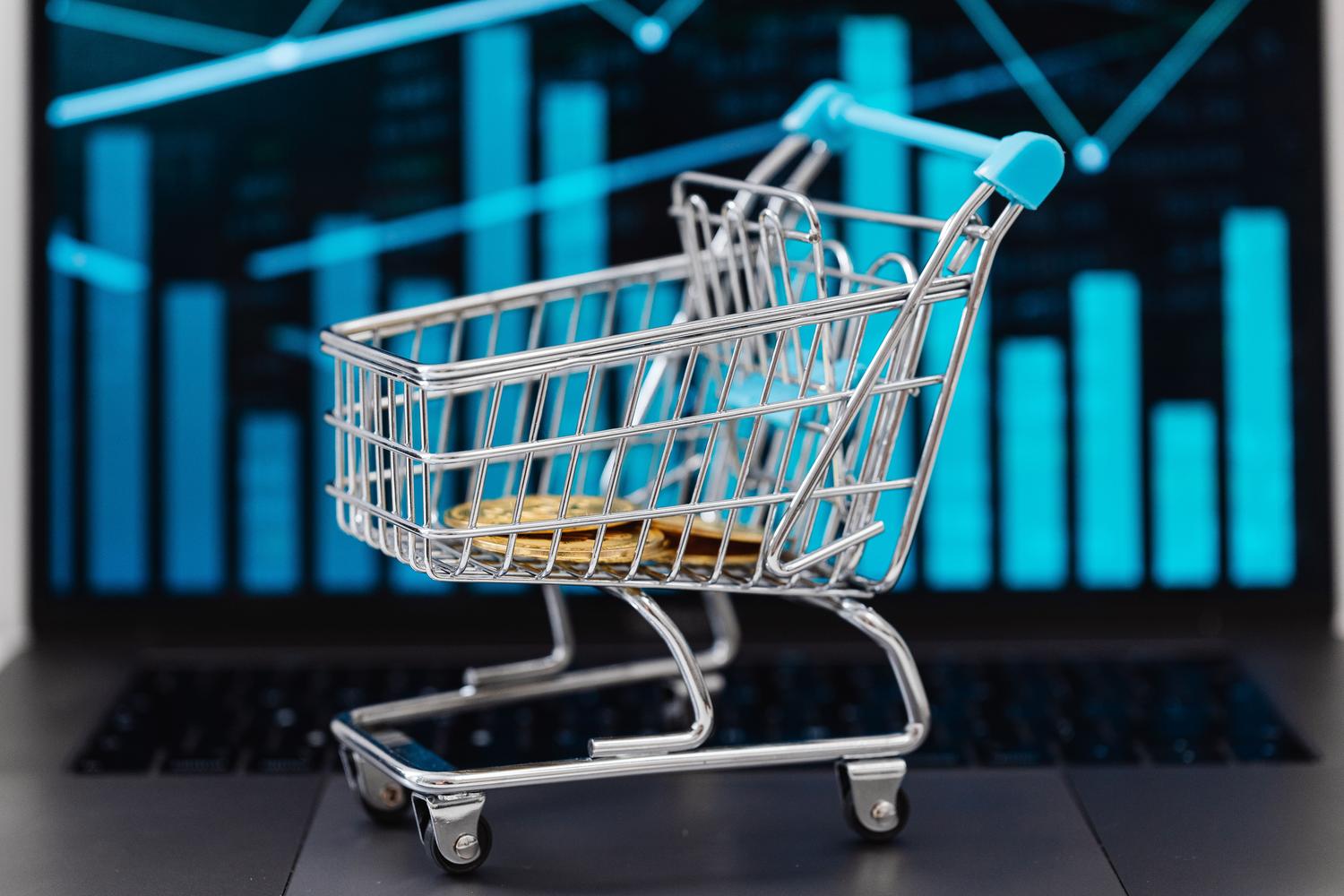 Miniature metal shopping cart with gold coins resting on a laptop keyboard with financial bar charts in the background, representing profitability through price monitoring.