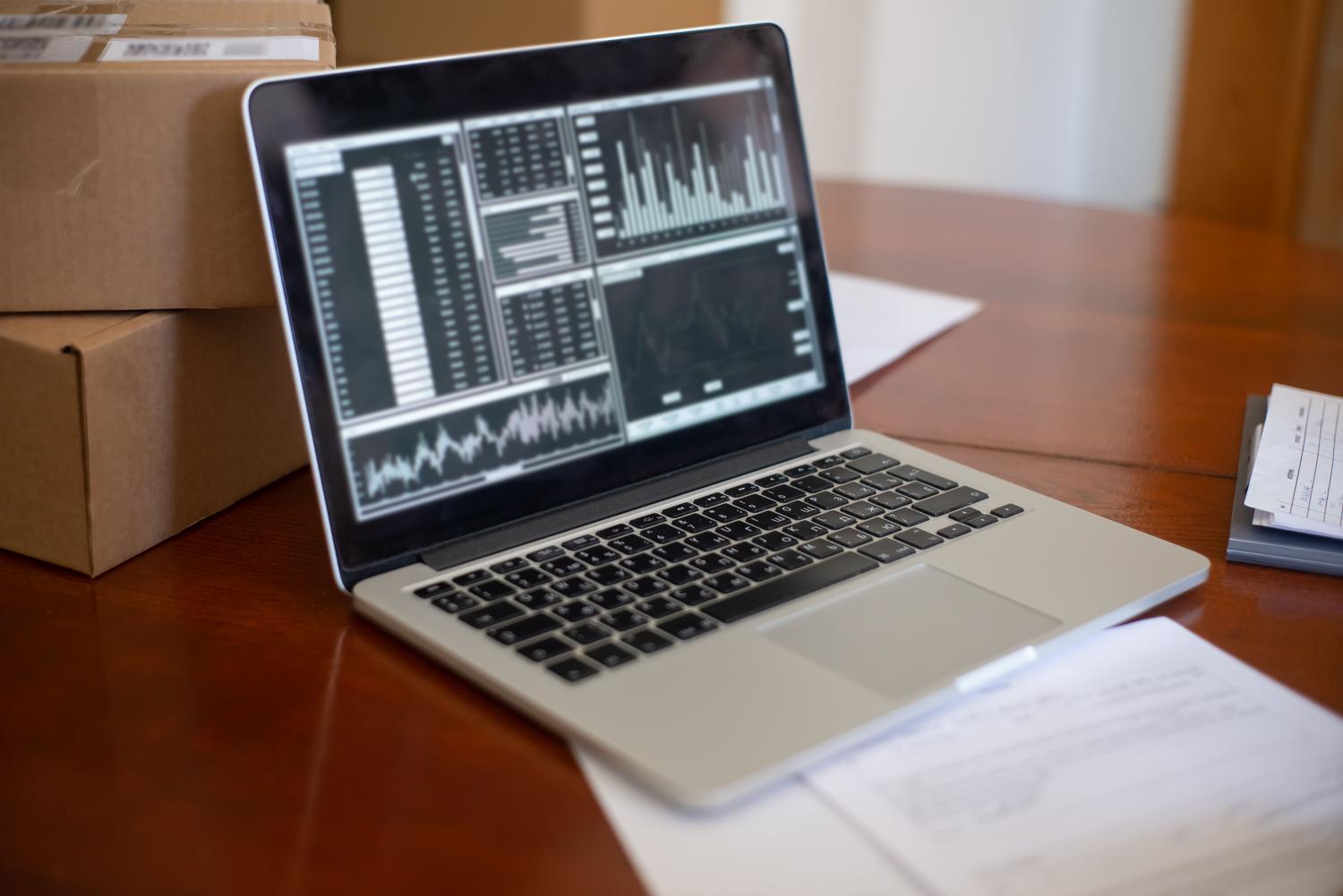 Laptop displaying an e-commerce analytics dashboard with price tracking graphs next to a stack of shipping boxes on a wooden desk, connecting data extraction with logistics.