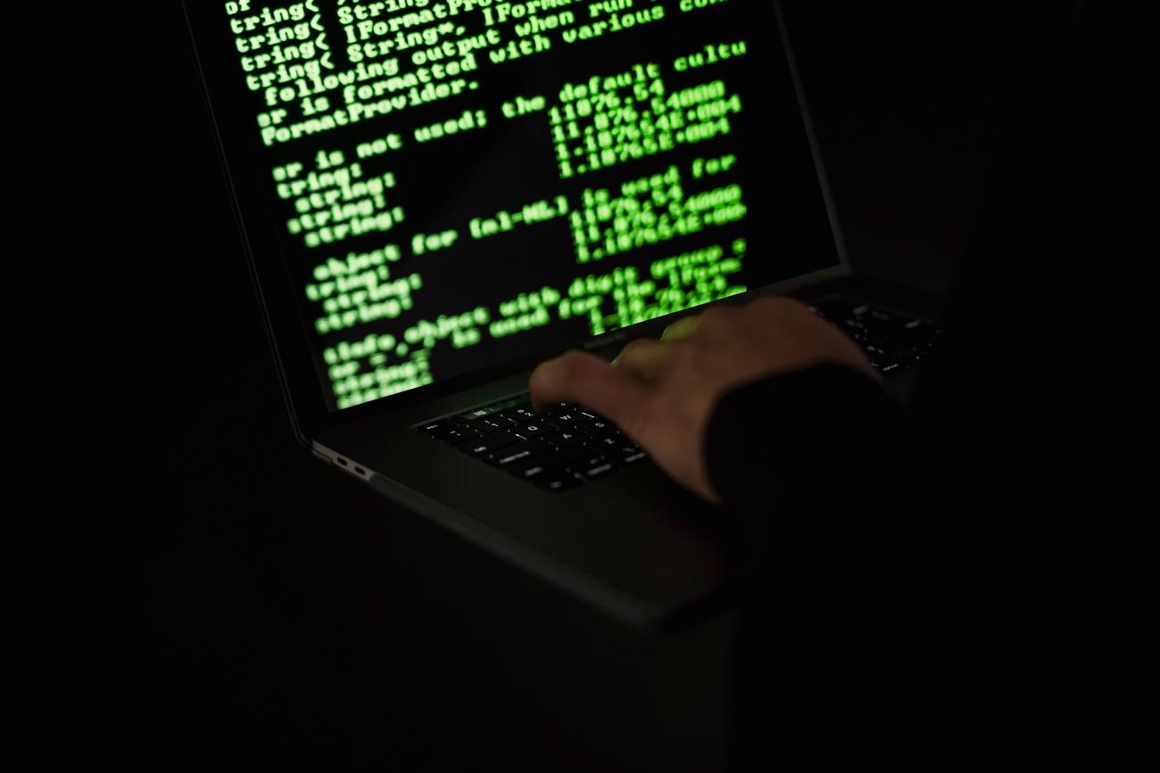 Persona encapuchada escribiendo código en un portátil en la oscuridad, representando un ataque de malware.