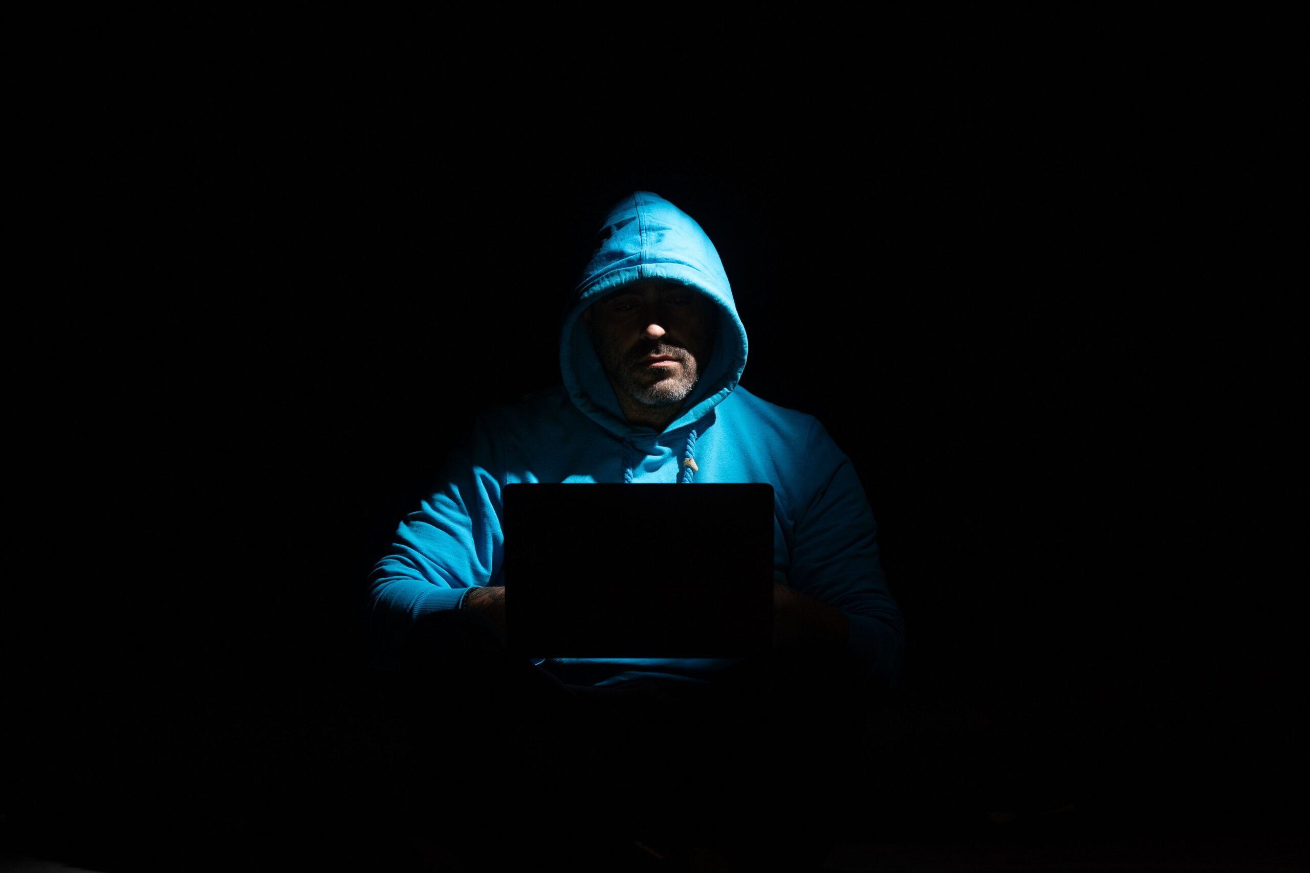 Persona anónima con capucha usando un portátil en la oscuridad, representando la amenaza invisible de los espías digitales.