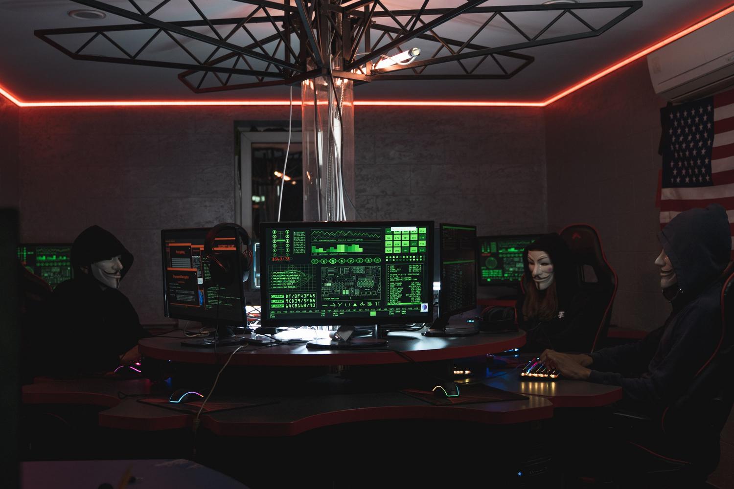 Grupo de ciberdelincuentes com máscaras en una habitación oscura llena de monitores, representando los ataques sofisticados.