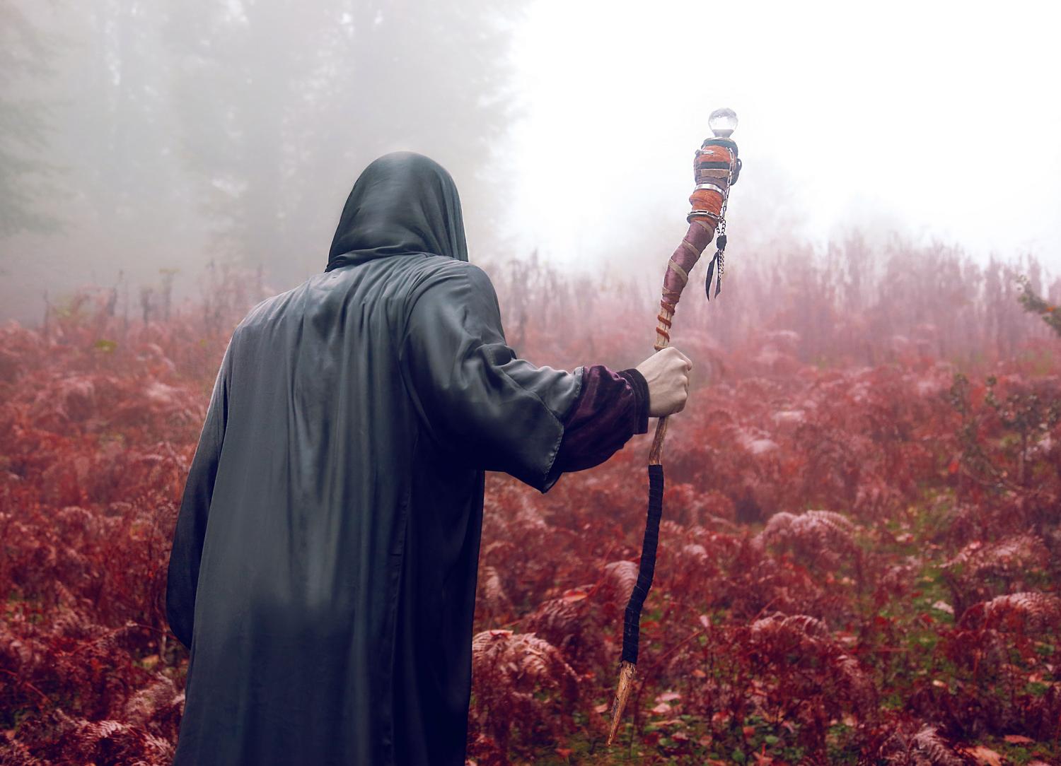 Figura encapuchada com báculo mágico en un bosque brumoso, evocando el rol de bruja de la protagonista