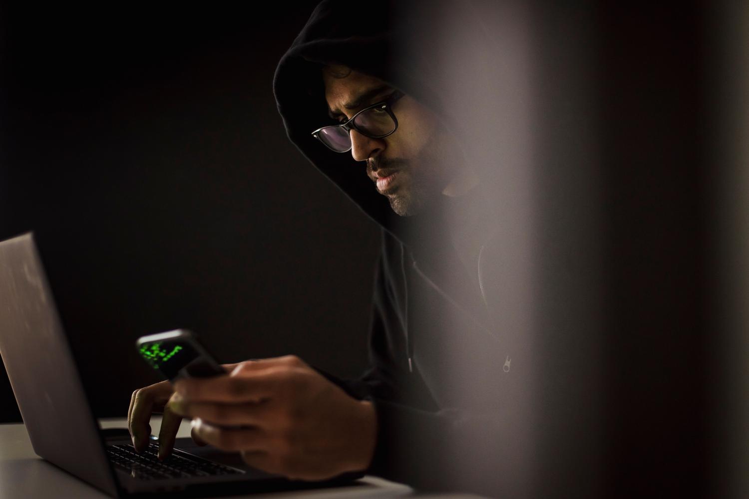 Representación de un hacker en un entorno oscuro operando un ordenador y un móvil, simbolizando el robo de cuentas.