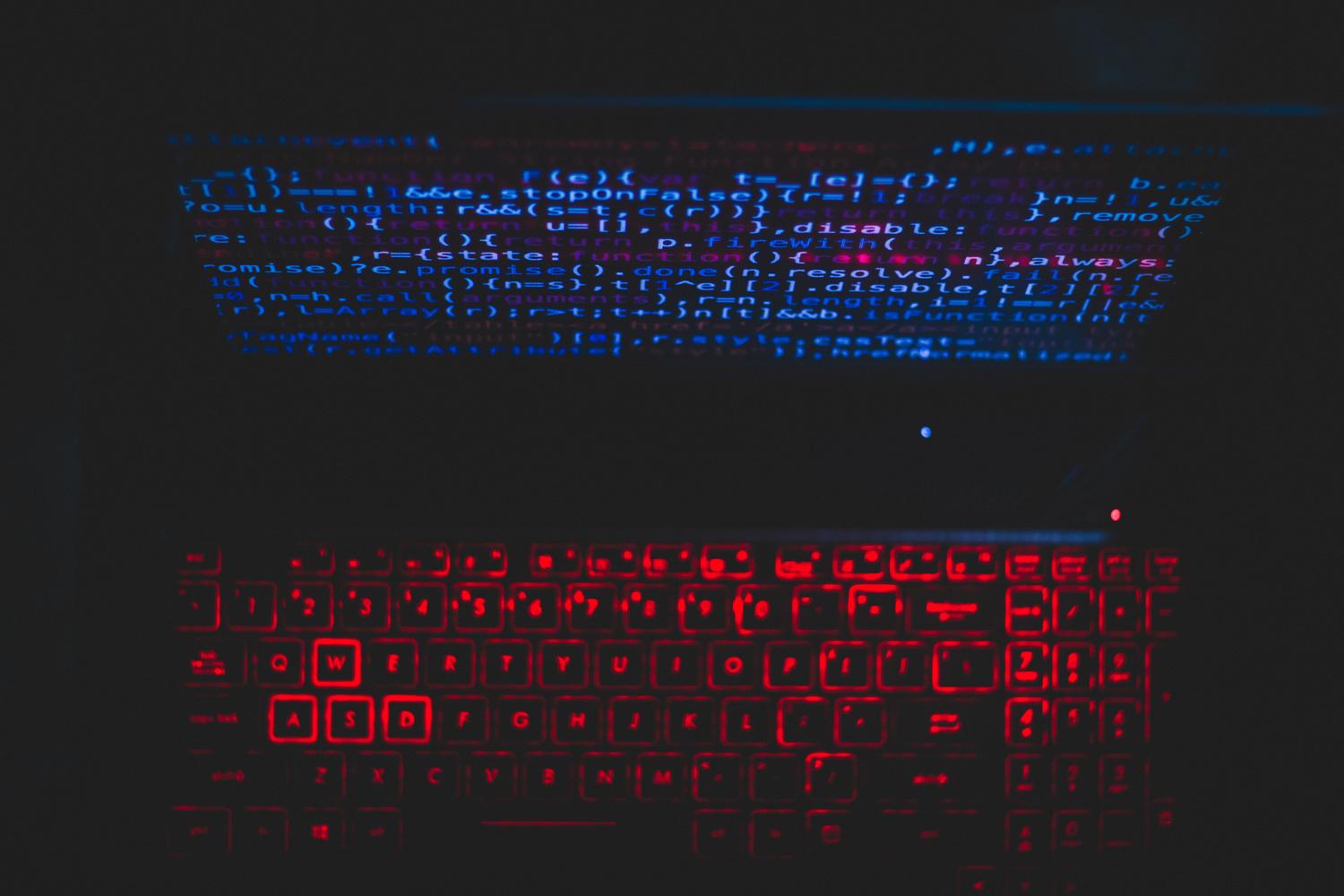Laptop en entorno oscuro con teclado iluminado en rojo y código en pantalla, simbolizando ciberataques y programación maliciosa.