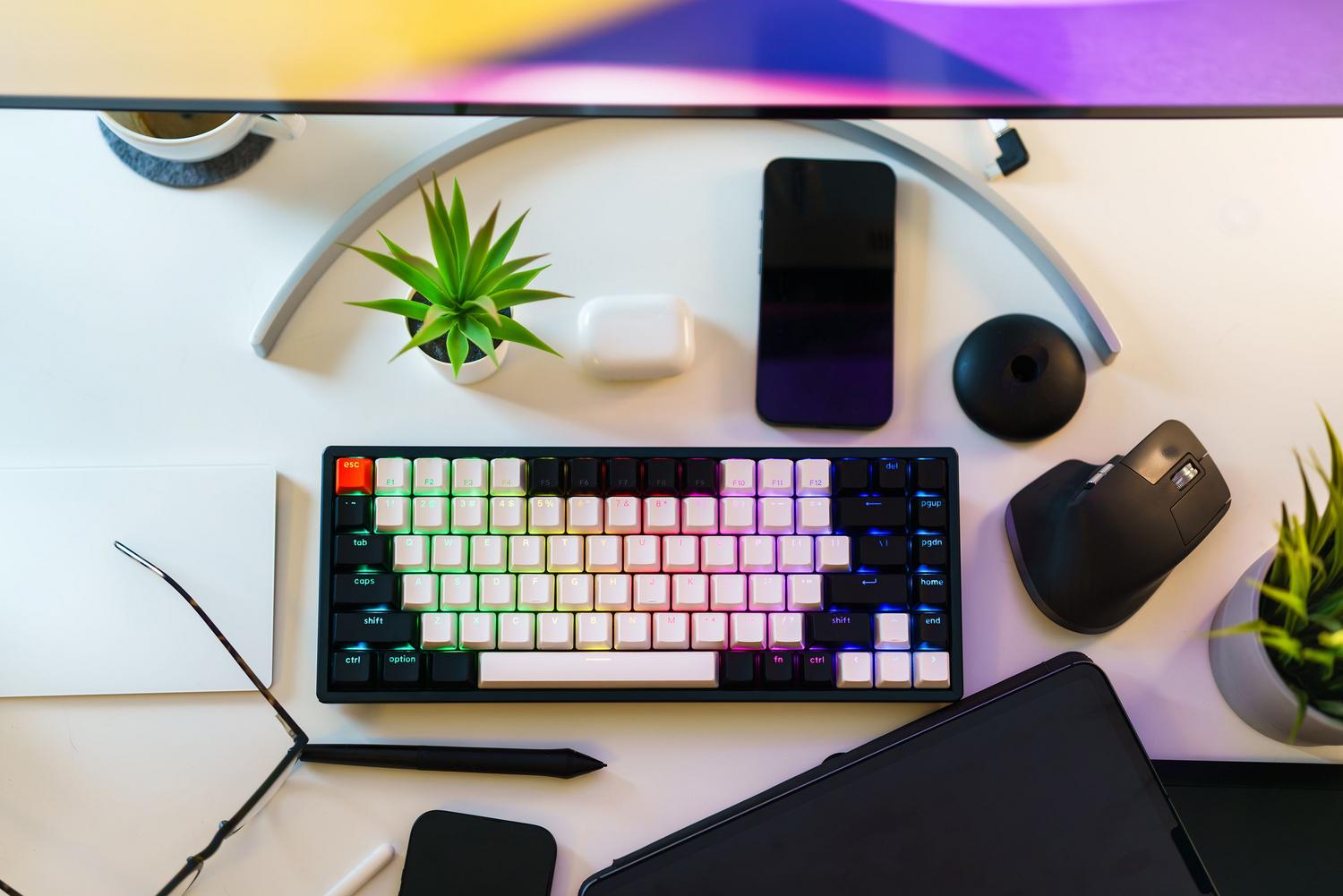 Escritorio blanco y limpio con teclado mecánico RGB, ratón ergonómico vertical y plantas, enfocado en la productividad.