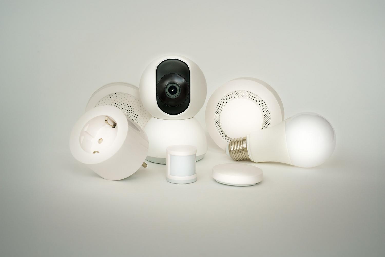 Conjunto de dispositivos IoT para el hogar, como cámaras de seguridad, enchufes inteligentes y bombillas LED sobre fondo blanco.