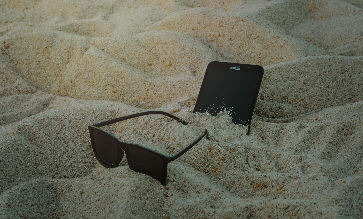 Teléfono móvil y gafas de sol sobre la arena de la playa, ilustrando la exposición al sol y superficies que retienen el calor.